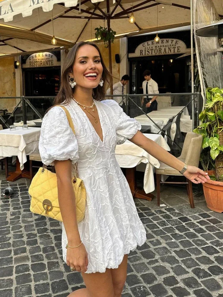 Model draagt een luxe geborduurde bloemenjurk met pofmouwen, een diepe V-hals en een speelse ballonzoom. De jurk heeft verfijnde borduurdetails en een elegante pasvorm, perfect voor zomerse dagen en feestelijke gelegenheden