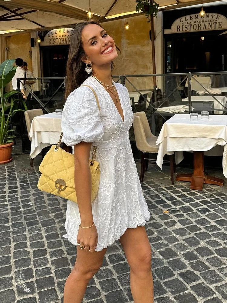 Model draagt een luxe geborduurde bloemenjurk met pofmouwen, een diepe V-hals en een speelse ballonzoom. De jurk heeft verfijnde borduurdetails en een elegante pasvorm, perfect voor zomerse dagen en feestelijke gelegenheden