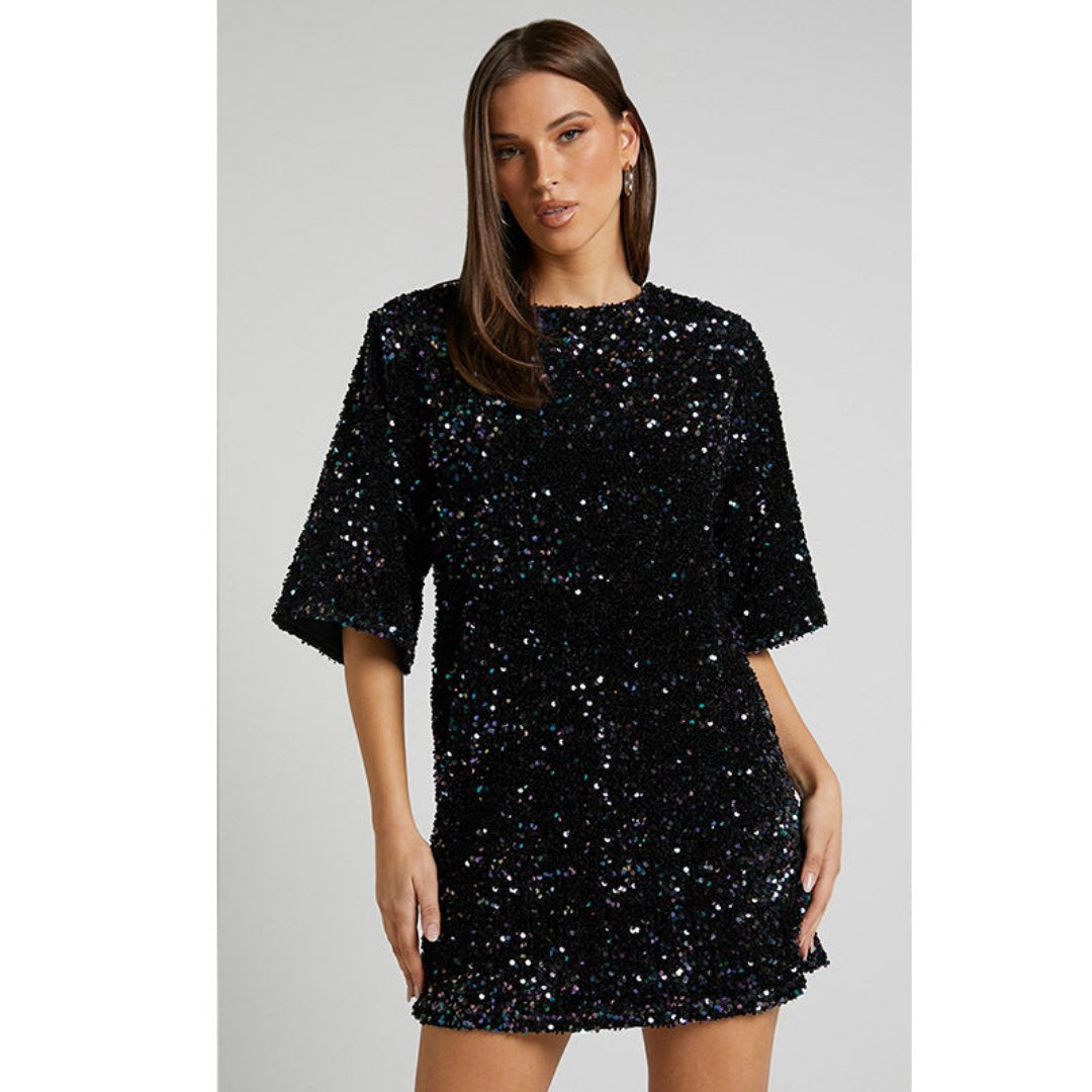Model draagt een luxe mini-jurk met pailletten in roze en zwart, met korte mouwen en open rug met strikdetail – perfect voor feestelijke gelegenheden of avondjes uit.