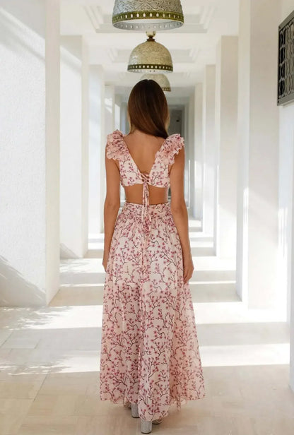 Vrouw draagt een romantische lange zomerjurk met roze bloemenprint, diepe V-hals en hoge split – perfect voor bruiloften of vakanties.