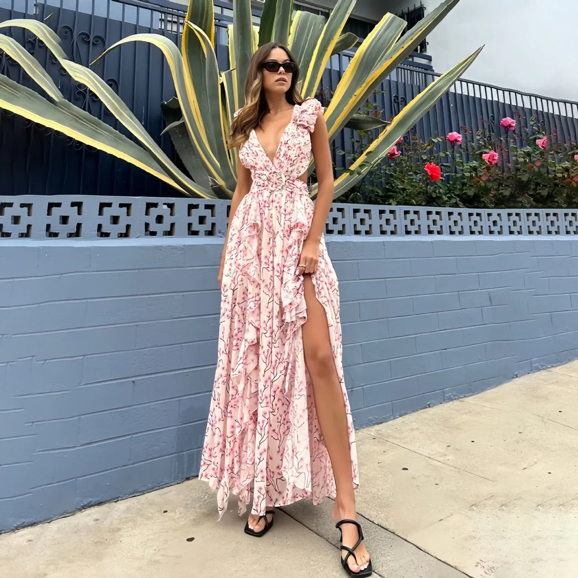 Vrouw draagt een romantische lange zomerjurk met roze bloemenprint, diepe V-hals en hoge split – perfect voor bruiloften of vakanties.