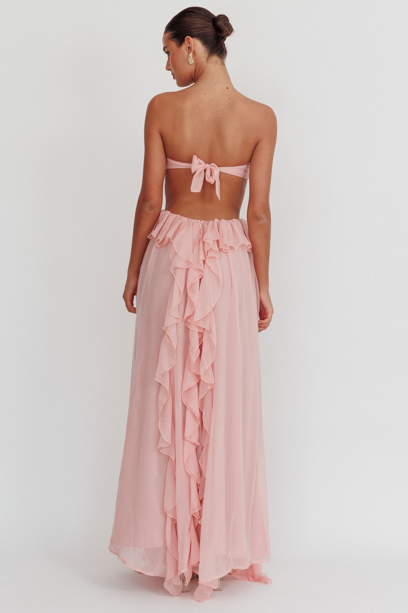 Model draagt een blush roze maxi jurk met ruches, strapless top en open rug, perfect voor zomerse evenementen.