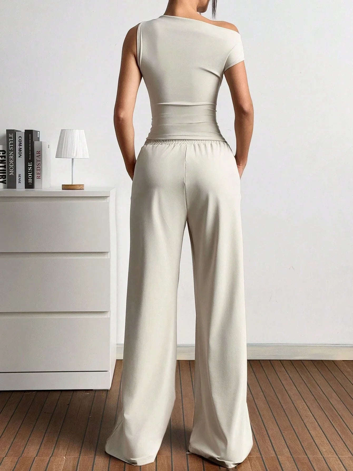 Dames in one-shoulder broekenset met wijdvallende broek, witte sneakers en rode handtas, poserend in een minimalistisch interieur.
