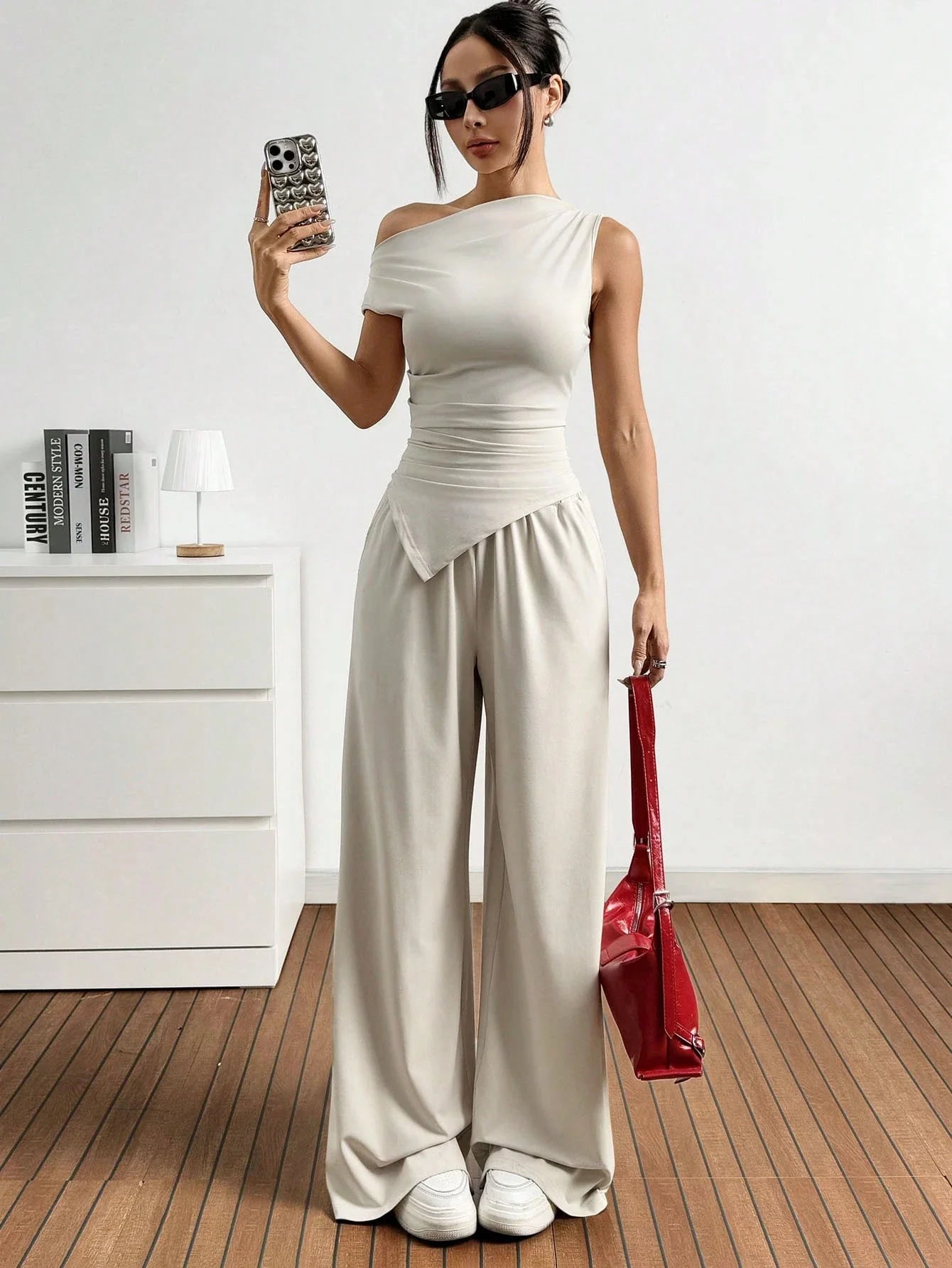 Dames in one-shoulder broekenset met wijdvallende broek, witte sneakers en rode handtas, poserend in een minimalistisch interieur.
