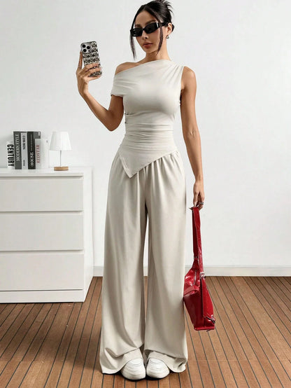 Dames in one-shoulder broekenset met wijdvallende broek, witte sneakers en rode handtas, poserend in een minimalistisch interieur.
