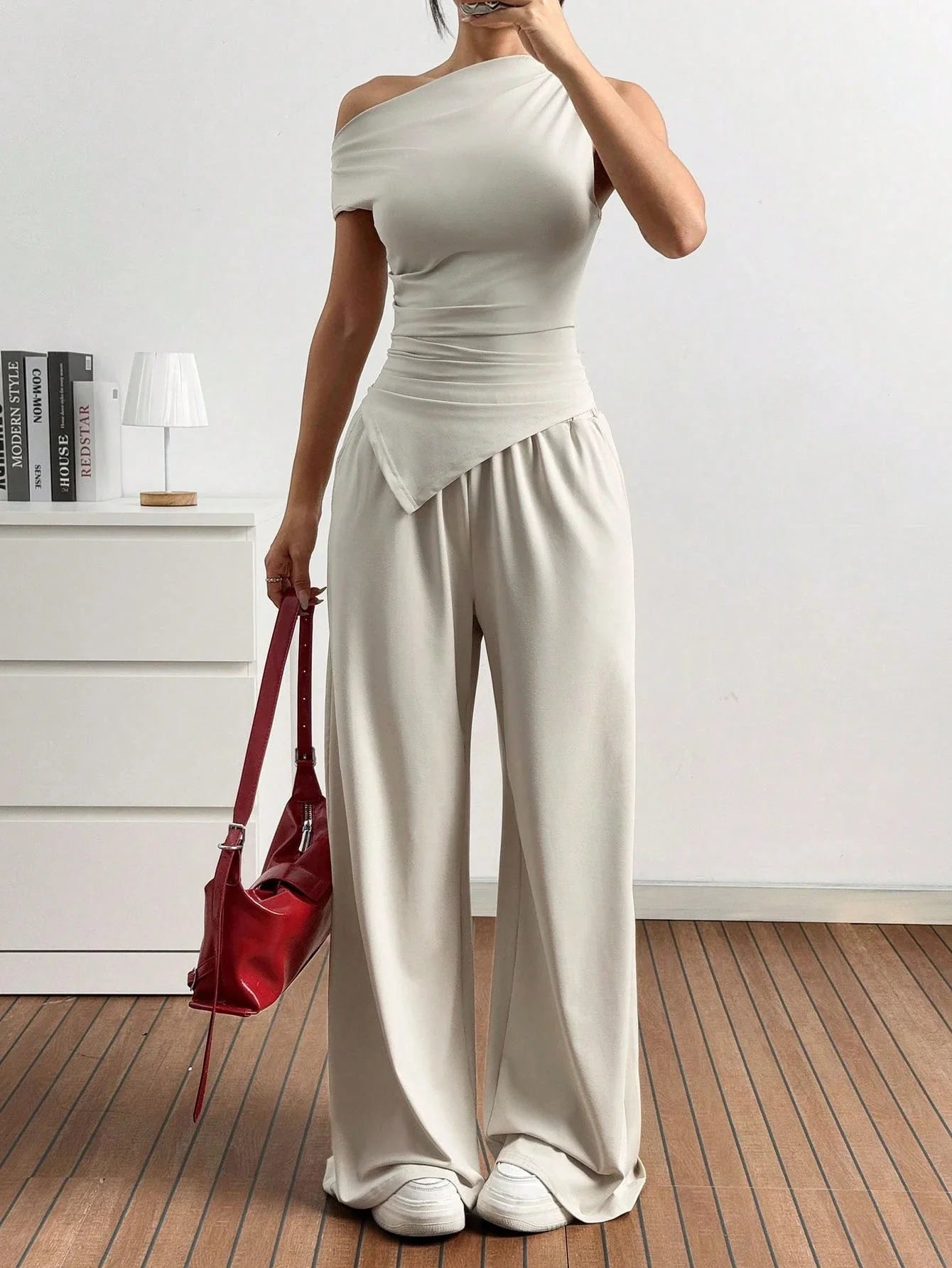 Dames in one-shoulder broekenset met wijdvallende broek, witte sneakers en rode handtas, poserend in een minimalistisch interieur.
