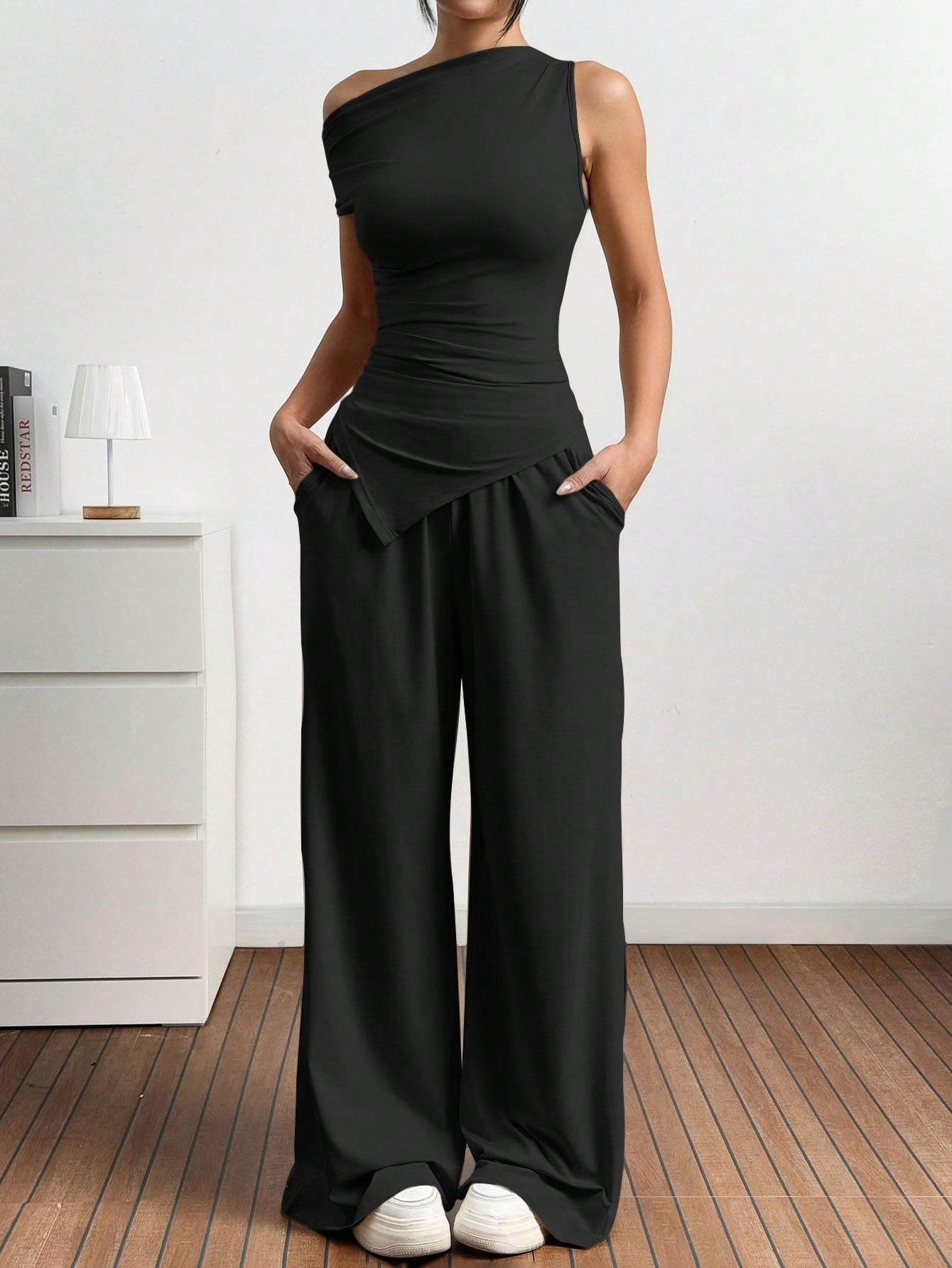Dames in one-shoulder broekenset met wijdvallende broek, witte sneakers en rode handtas, poserend in een minimalistisch interieur.
