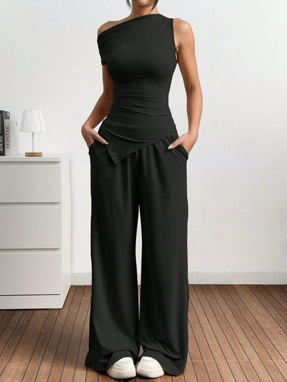 Dames in one-shoulder broekenset met wijdvallende broek, witte sneakers en rode handtas, poserend in een minimalistisch interieur.

